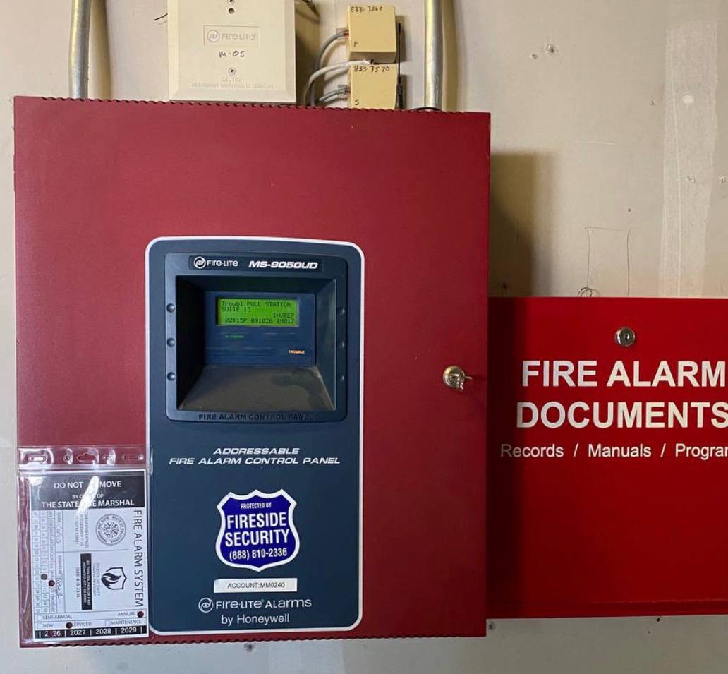 Fire-Lite MS-9050UD commercial fire alarm control panel with Fireside Security monitoring badge, California inspection tag, and red fire alarm documents box